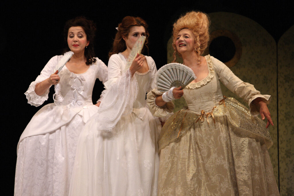 Tri glumice u raskošnim povijesnim kostimima iz 18. stoljeća stoje na pozornici. Sve nose duge raskošne haljine u bijelim i zlatnim tonovima te drže lepeze u rukama. Izrazima lica djeluju kao da su u živahnom razgovoru.