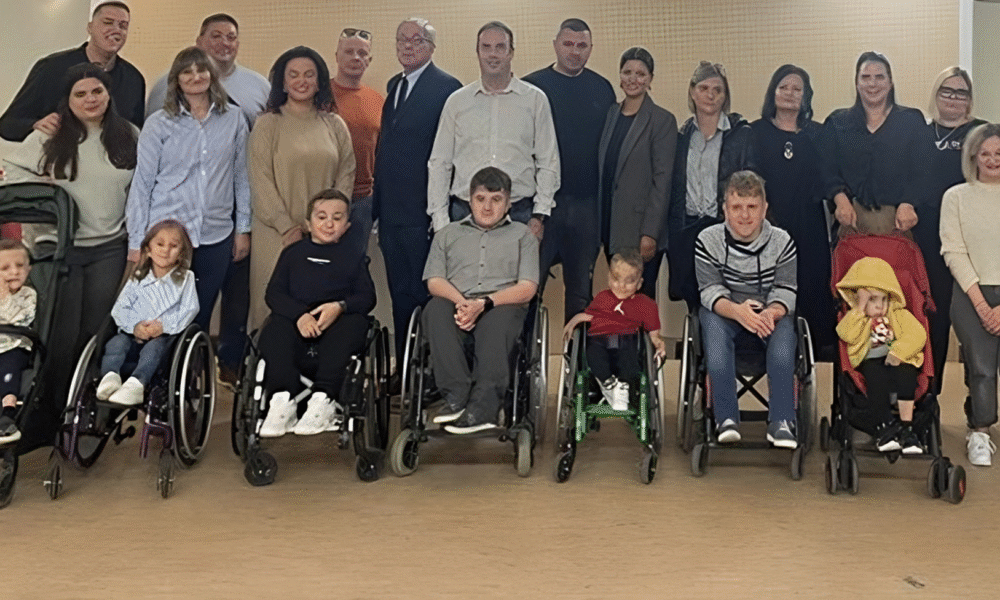 Na fotografiji je prikazana skupina ljudi okupljena u zatvorenom prostoru, vjerojatno povodom društvenog ili zajedničkog događanja. U prvom redu sjede djeca i odrasli koji koriste invalidska kolica ili kolica za djecu, dok iza njih stoje ostali sudionici – raznolika skupina različitih dobi. Svi su okrenuti prema kameri i poziraju s osmijehom, stvarajući dojam zajedništva, podrške i inkluzije. Atmosfera je topla i prijateljska, a postav fotografije naglašava ravnopravnost i vidljivost osoba s invaliditetom u društvenim okruženjima. Ova slika snažno prenosi poruku prihvaćanja, solidarnosti i važnosti zajedničkih trenutaka.