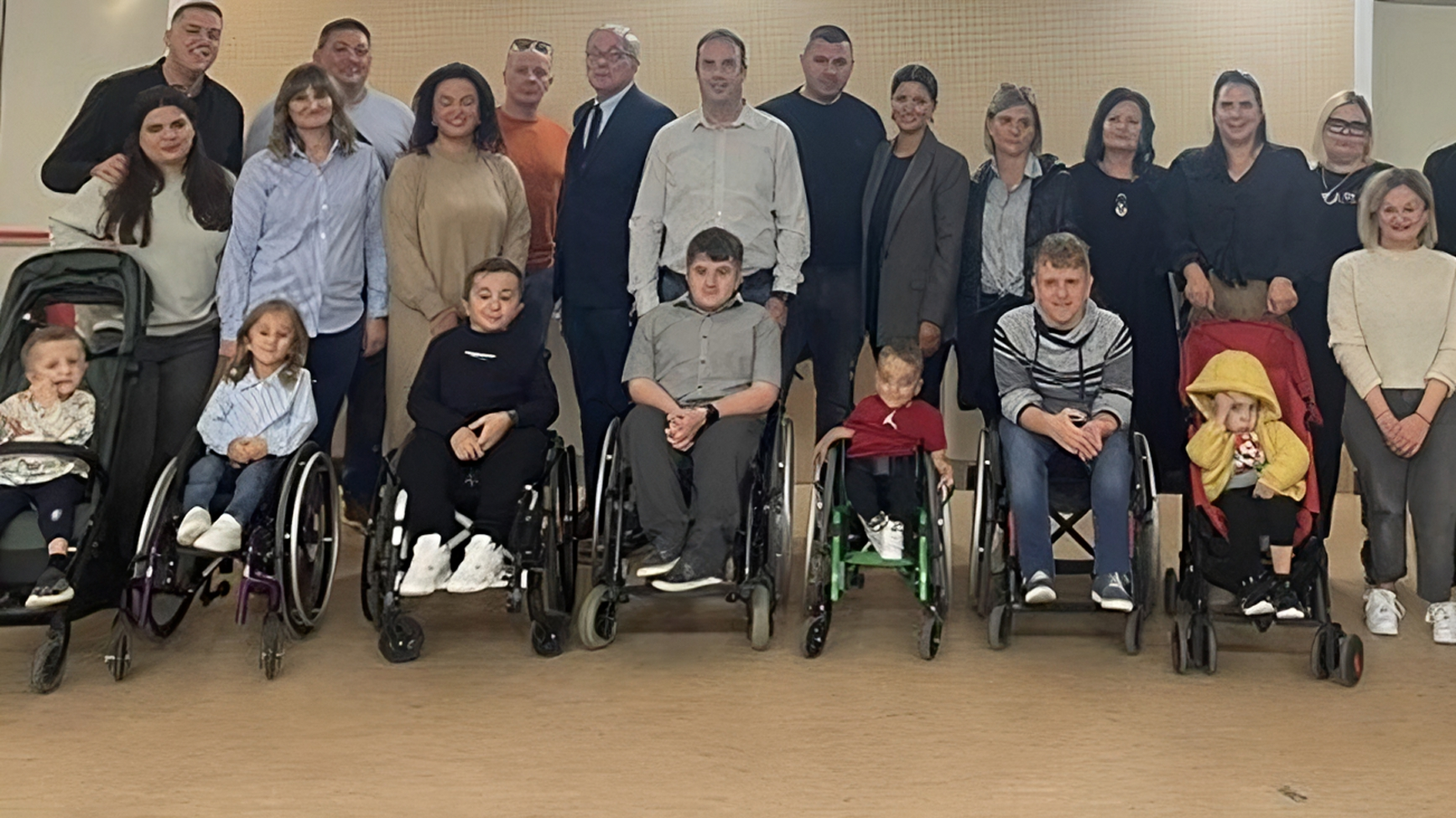 Na fotografiji je prikazana skupina ljudi okupljena u zatvorenom prostoru, vjerojatno povodom društvenog ili zajedničkog događanja. U prvom redu sjede djeca i odrasli koji koriste invalidska kolica ili kolica za djecu, dok iza njih stoje ostali sudionici – raznolika skupina različitih dobi. Svi su okrenuti prema kameri i poziraju s osmijehom, stvarajući dojam zajedništva, podrške i inkluzije. Atmosfera je topla i prijateljska, a postav fotografije naglašava ravnopravnost i vidljivost osoba s invaliditetom u društvenim okruženjima. Ova slika snažno prenosi poruku prihvaćanja, solidarnosti i važnosti zajedničkih trenutaka.