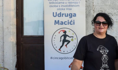 Na fotografiji se nalazi žena koja stoji iza stola prekrivenog promotivnim materijalima. Nosi sunčane naočale i crnu majicu s crtežom simpatičnog lika i natpisom „Macići“. Ispred nje je stol s natpisom „UDRUGA MACIĆI“, a iza nje se nalazi veliki banner s tekstom.