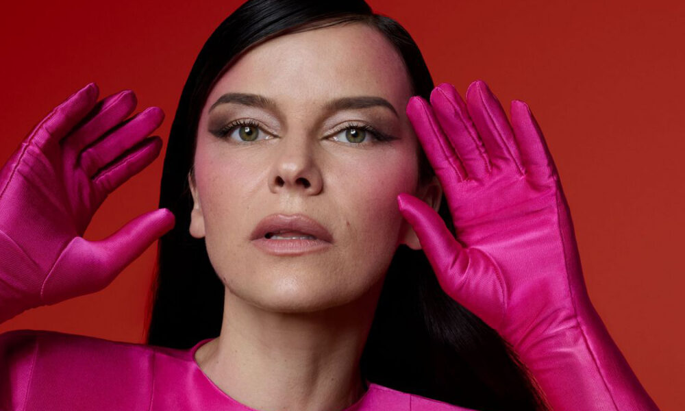 Portret žene ispred jarko crvene pozadine. Pozirala je frontalno, podignutih ruku uz lice, na kojima nosi sjajne, ružičaste rukavice. Odjevena je u ružičasti gornji dio, a kosa joj je tamna i zalizana unatrag. Pogled joj je usmjeren prema naprijed, uz neutralan izraz lica.