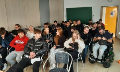 Na fotografiji se nalazi učionica ili seminar sala ispunjena ljudima, najvjerojatnije studentima ili sudionicima nekog predavanja ili radionice. Prostorija ima svijetle zidove, ormariće uz zid, zelenu školsku ploču i žuta vrata na kojima je zalijepljen plakat. Većina osoba sjedi u redovima stolica, neki koriste prijenosna računala ili mobitele, a jedna osoba je u invalidskim kolicima. Atmosfera djeluje koncentrirano i angažirano, što sugerira da se radi o edukativnom ili informativnom događaju.