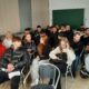Na fotografiji se nalazi učionica ili seminar sala ispunjena ljudima, najvjerojatnije studentima ili sudionicima nekog predavanja ili radionice. Prostorija ima svijetle zidove, ormariće uz zid, zelenu školsku ploču i žuta vrata na kojima je zalijepljen plakat. Većina osoba sjedi u redovima stolica, neki koriste prijenosna računala ili mobitele, a jedna osoba je u invalidskim kolicima. Atmosfera djeluje koncentrirano i angažirano, što sugerira da se radi o edukativnom ili informativnom događaju.