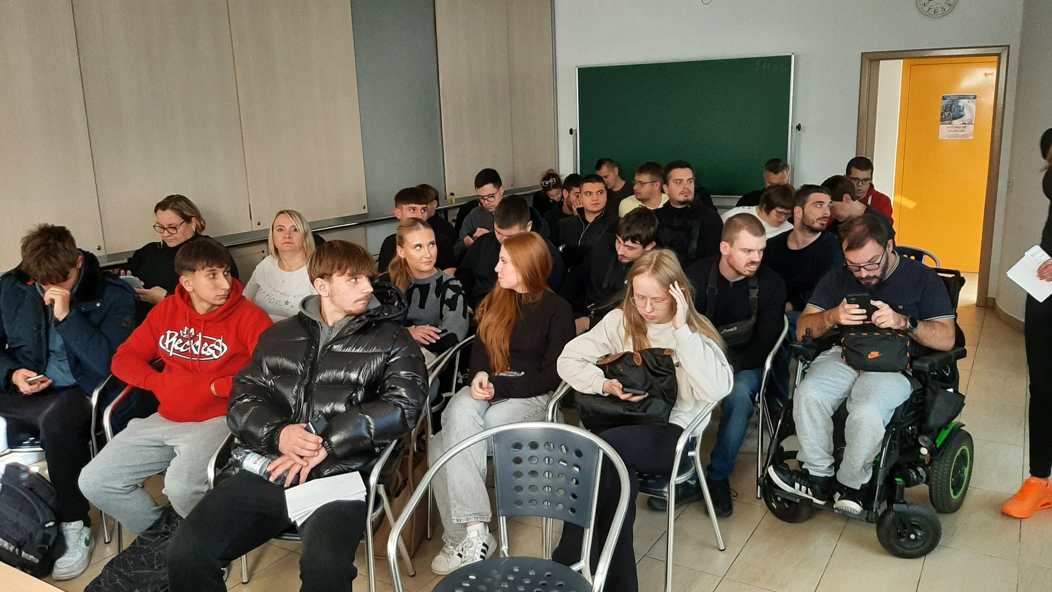 Na fotografiji se nalazi učionica ili seminar sala ispunjena ljudima, najvjerojatnije studentima ili sudionicima nekog predavanja ili radionice. Prostorija ima svijetle zidove, ormariće uz zid, zelenu školsku ploču i žuta vrata na kojima je zalijepljen plakat. Većina osoba sjedi u redovima stolica, neki koriste prijenosna računala ili mobitele, a jedna osoba je u invalidskim kolicima. Atmosfera djeluje koncentrirano i angažirano, što sugerira da se radi o edukativnom ili informativnom događaju.
