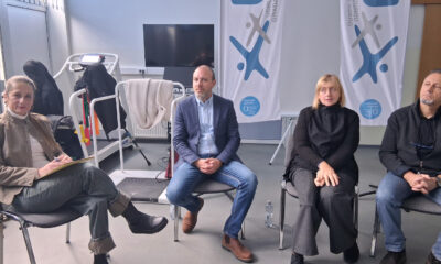 Četvero odraslih osoba sjedi u krugu u svijetloj dvorani, kao na sastanku ili radionici. S lijeva na desno: žena s bilježnicom u ruci, muškarac u sakou i trapericama, žena u crnoj odjeći te muškarac u tamnoj jakni. Iza njih se vide sprave za rehabilitaciju, televizor i dvije velike bijele zastave s logom udruge. Atmosfera je mirna i profesionalna.