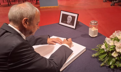 Muškarac u odijelu upisuje se u knjigu žalosti na stolu prekrivenom tamnim stolnjakom; pored njega su fotografija preminule osobe u okviru, upaljena svijeća i cvjetni aranžman s bijelim orhidejama.