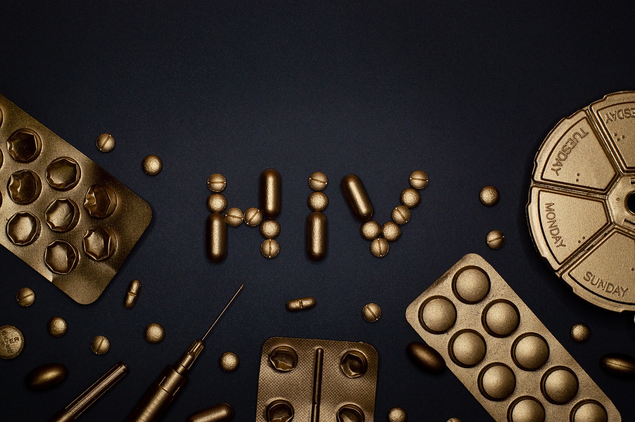 Fotografija prikazuje snažnu i simboličnu kompoziciju posvećenu svijesti o HIV-u i važnosti liječenja. Na tamnoj pozadini ističu se medicinski predmeti zlatne boje — pilule, tablete, blister pakiranja, štrcaljka, olovkasti predmet i okrugli organizator za lijekove s oznakama dana u tjednu (ponedjeljak, utorak, nedjelja). U središtu fotografije, pilule su pažljivo posložene tako da tvore natpis „HIV“, čime se vizualno naglašava tema bolesti i terapije. Zlatna boja lijekova simbolizira vrijednost i nadu koju donosi pristup medicini, dok tamna pozadina pojačava kontrast i dramatičnost prizora. Ova fotografija prenosi poruku o važnosti redovitog uzimanja terapije, dostupnosti lijekova i borbi protiv stigme povezane s HIV-om. Vizualno je upečatljiva, emocionalno snažna i edukativna.