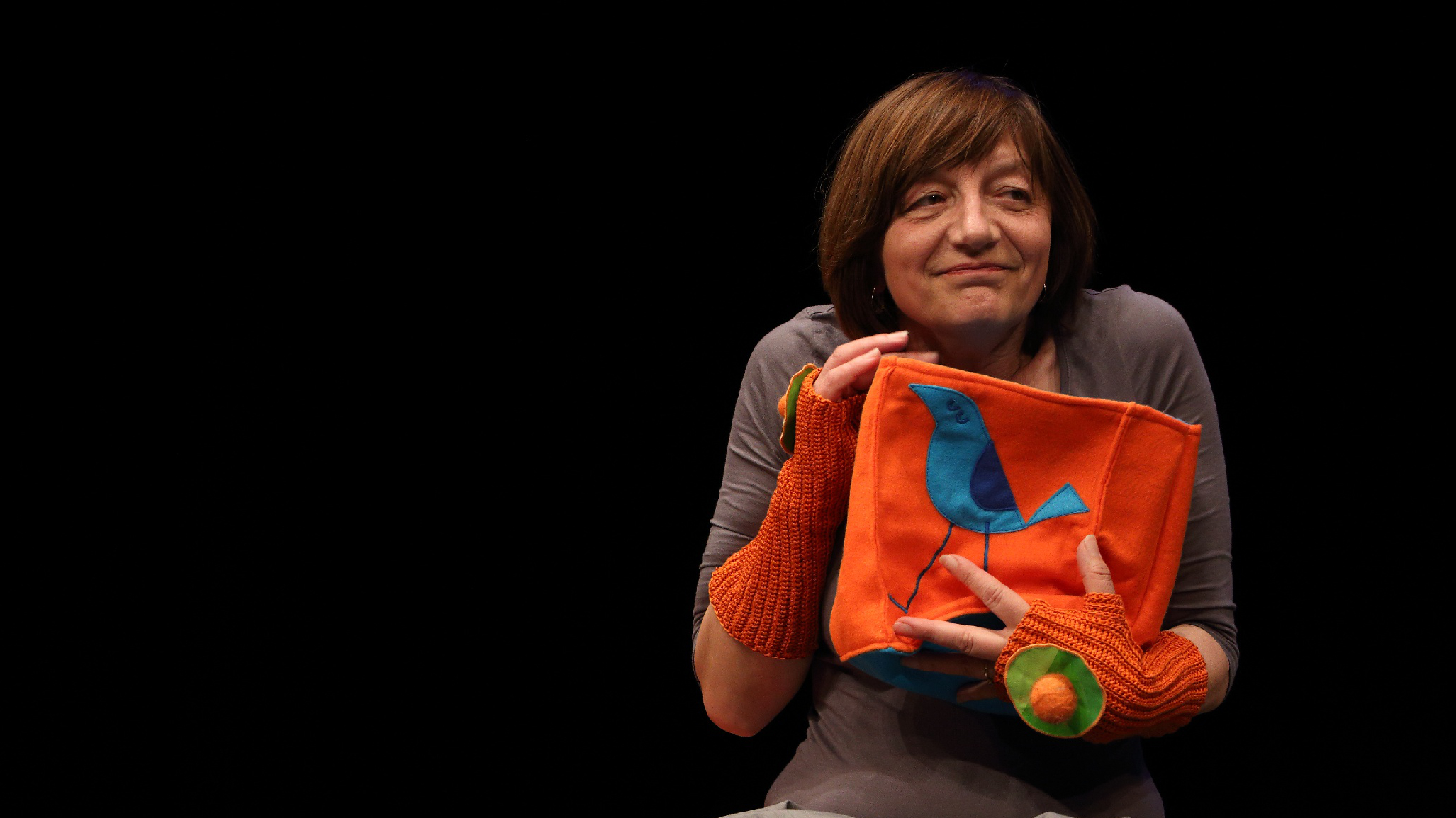 Glumica sjedi na narančastoj klupi i drži narančastu torbicu s plavom pticom, nasmiješena i okrenuta blago u stranu. Pozadina je crna, bez dodatnih elemenata scenografije.