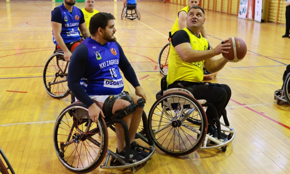 Fotografija prikazuje utakmicu košarke u kolicima. Jedna momčad je u žutim, a druga u plavim dresovima, a na slici igrač u žitom drži loptu dok je igrač u plavom dresu pokraj njega.