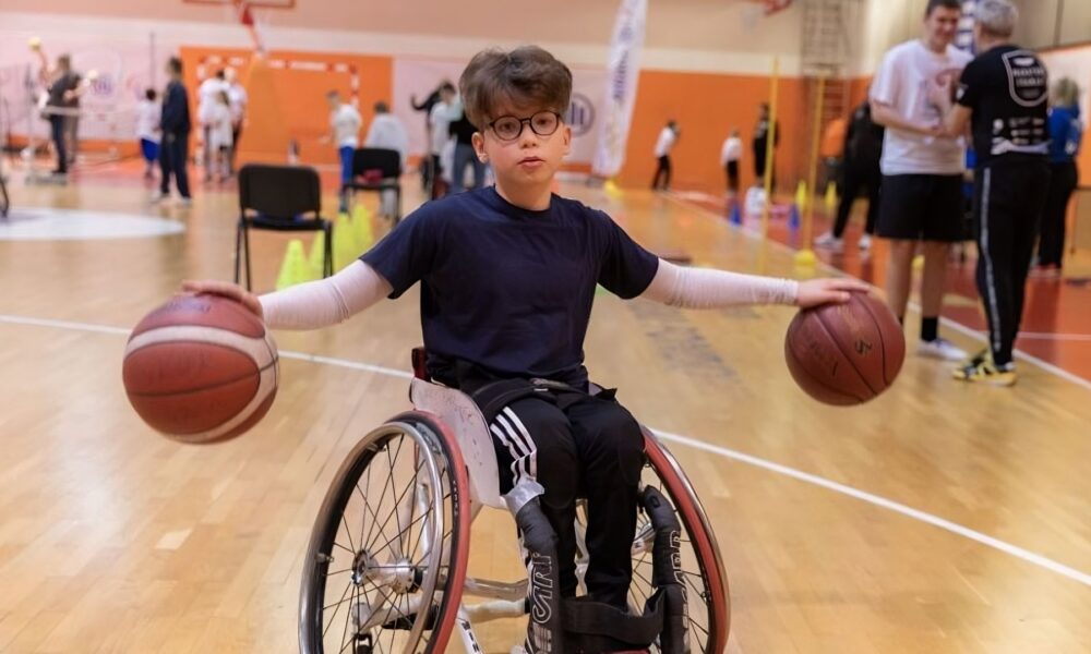 Dječak u invalidskim kolicima nalazi se u sportskoj dvorani i drži dvije košarkaške lopte – jednu u svakoj ruci – s raširenim rukama. Na sebi ima tamnoplavu majicu, crne trenirke s bijelim prugama i naočale. U pozadini se vide druga djeca, treneri i sportska oprema, što sugerira da je u tijeku sportski trening ili radionica inkluzivne košarke.