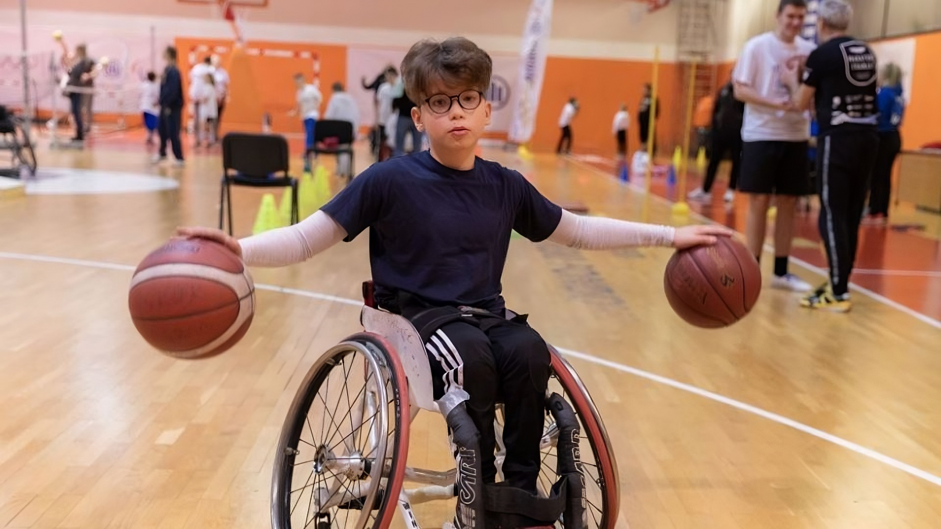 Dječak u invalidskim kolicima nalazi se u sportskoj dvorani i drži dvije košarkaške lopte – jednu u svakoj ruci – s raširenim rukama. Na sebi ima tamnoplavu majicu, crne trenirke s bijelim prugama i naočale. U pozadini se vide druga djeca, treneri i sportska oprema, što sugerira da je u tijeku sportski trening ili radionica inkluzivne košarke.