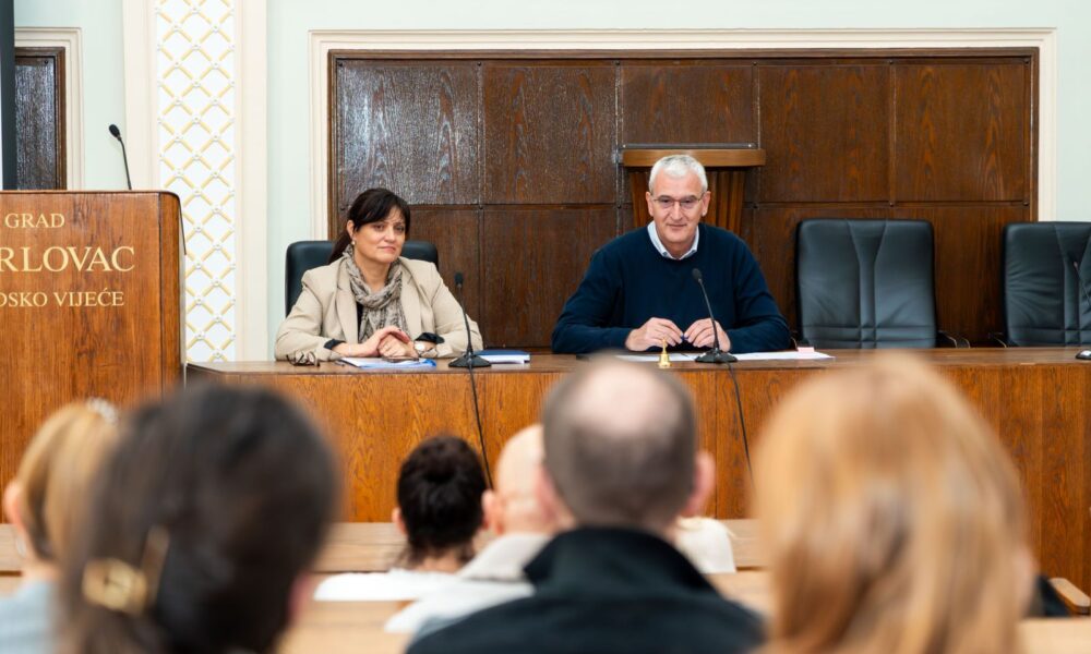 Na fotografiji je prikazana službena dvorana u kojoj se održava sastanak ili sjednica. U prvom planu vidi se publika okrenuta prema govornicima, dok su u drugom planu za velikim drvenim stolom smještene dvije osobe. Sjednu za stolom, ispred njih se nalaze mikrofoni i bilješke, što upućuje na to da vode sastanak ili iznose informacije prisutnima. Osoba s lijeve strane nosi svijetli sako i šal, te izgleda smireno i pažljivo prati zbivanje u dvorani. Osoba s desne strane odjevena je u tamniju odjeću i djeluje kao da se obraća prisutnima, s blagim osmijehom i rukama položenima na stol. Iza njih se nalaze drvene zidne obloge i velika govornica s natpisom koji upućuje na to da je riječ o prostoru lokalne vlasti. Atmosfera djeluje profesionalno i službeno, a izrazima lica prisutnih stječe se dojam da je sastanak važan i usmjeren na ozbiljne teme.