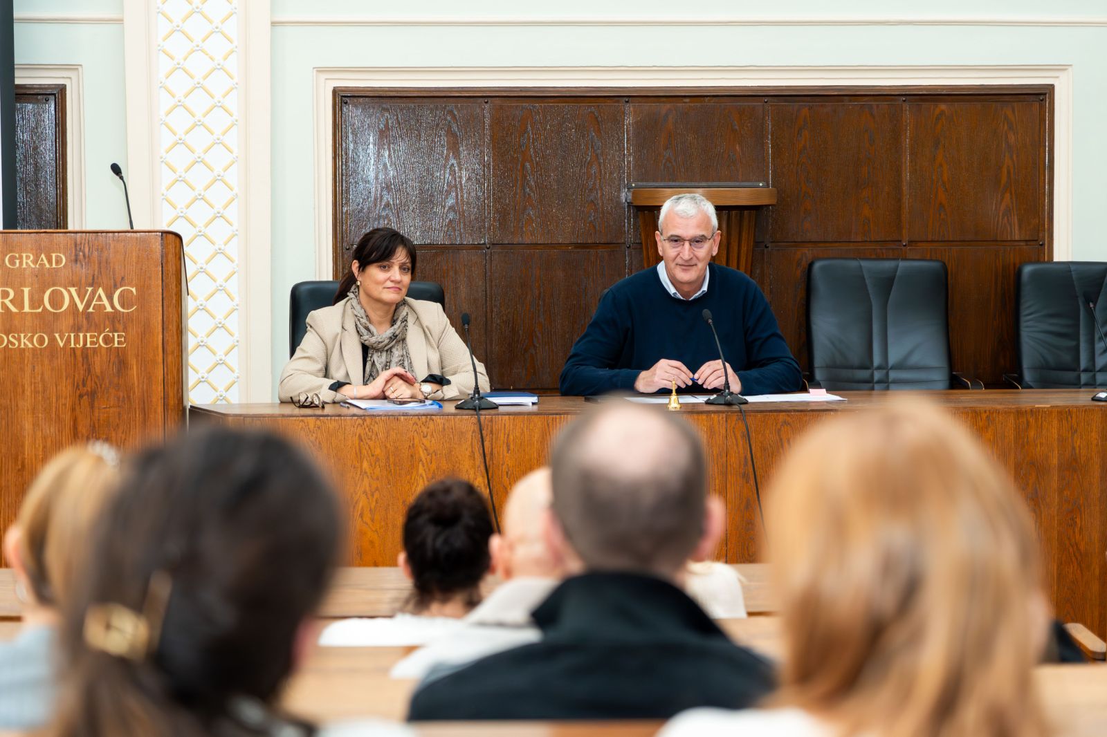 Na fotografiji je prikazana službena dvorana u kojoj se održava sastanak ili sjednica. U prvom planu vidi se publika okrenuta prema govornicima, dok su u drugom planu za velikim drvenim stolom smještene dvije osobe. Sjednu za stolom, ispred njih se nalaze mikrofoni i bilješke, što upućuje na to da vode sastanak ili iznose informacije prisutnima. Osoba s lijeve strane nosi svijetli sako i šal, te izgleda smireno i pažljivo prati zbivanje u dvorani. Osoba s desne strane odjevena je u tamniju odjeću i djeluje kao da se obraća prisutnima, s blagim osmijehom i rukama položenima na stol. Iza njih se nalaze drvene zidne obloge i velika govornica s natpisom koji upućuje na to da je riječ o prostoru lokalne vlasti. Atmosfera djeluje profesionalno i službeno, a izrazima lica prisutnih stječe se dojam da je sastanak važan i usmjeren na ozbiljne teme.