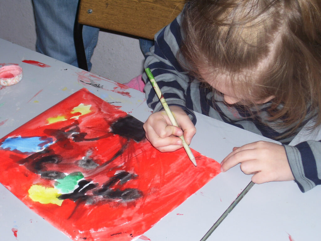 Djevojčica u prugastoj majici naginje se nad stol dok kistom oslikava papir živih boja, pretežno crvene, crne i plave.