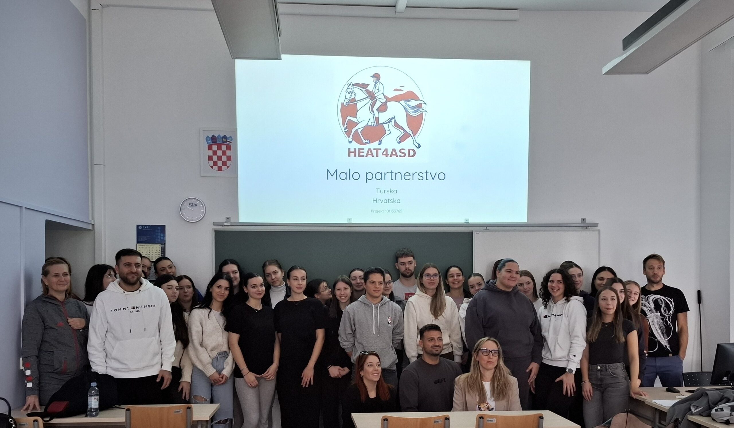 Na fotografiji se nalazi skupina ljudi, vjerojatno učenika i instruktora, okupljenih u učionici. Poziraju za zajedničku fotografiju ispred projekcijskog platna na kojem se prikazuje prezentacijski slajd. Na slajdu se nalazi logotip s prikazom osobe na konju i tekst "HEAT4ASD". Ispod logotipa piše: "Malo partnerstvo Turska Hrvatska Projekt 101135765" Ova oznaka upućuje na međunarodni projekt suradnje između Turske i Hrvatske, najvjerojatnije vezan uz obrazovanje ili istraživanje u okviru inicijative HEAT4ASD. U prostoriji se vidi hrvatski grb na zidu, sat i kalendar, što dodatno potvrđuje da se događaj odvija u Hrvatskoj.
