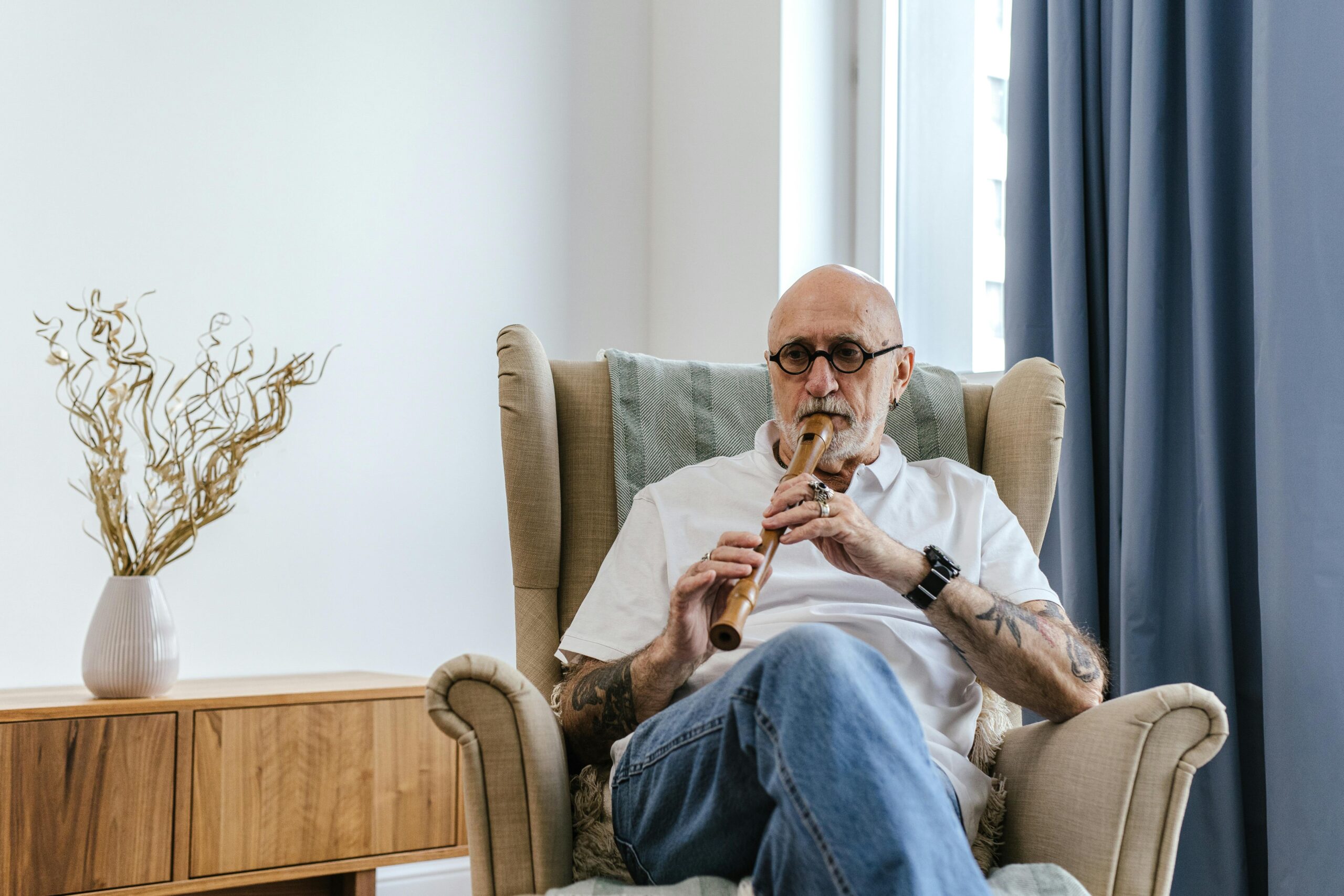 Fotografija prikazuje starijeg čovjeka kako sjedi na fotelji i svira flautu.