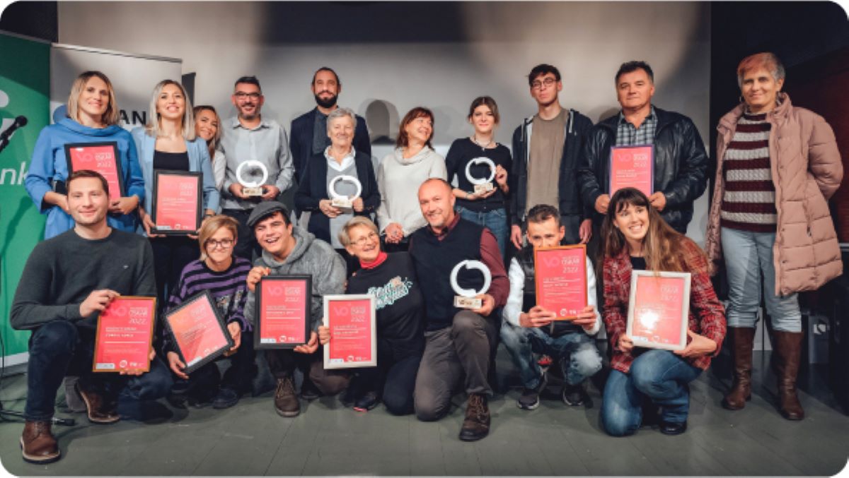 Na fotografiji se nalazi grupa od 17 osoba koje poziraju zajedno u zatvorenom prostoru, najvjerojatnije na pozornici ili u dvorani tijekom svečanosti dodjele nagrada. Većina sudionika drži crvene certifikate i okrugle trofeje, što upućuje na to da su primili priznanja za svoja postignuća. Atmosfera je svečana i vesela — svi izgledaju ponosno i zadovoljno.