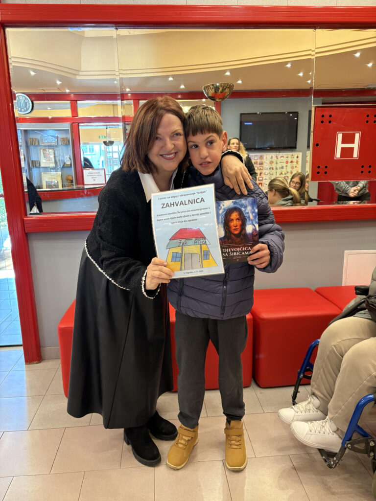 Žena i dječak stoje zagrljeni u prostoru s crvenim i zlatnim detaljima. Žena se nasmiješeno naginje prema dječaku i u rukama drži zahvalnicu s ilustracijom kuće. Dječak drži knjigu s naslovnicom na kojoj je prikazan lik djevojčice. U pozadini se vide djeca i odrasli, te dio invalidskih kolica.