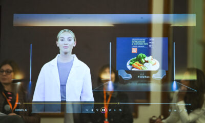 Fotografija prikazuje suvremeni konferencijski ambijent u kojem se odvija prezentacija uz pomoć hologramske ili proširene stvarnosti. Na središnjem ekranu projicirana je žena u bijelom laboratorijskom ogrtaču – vjerojatno virtualna govornica ili stručnjakinja koja sudjeluje na daljinu. Iza nje se nalazi digitalni prikaz tanjura s hranom izrađenom od šarenih građevnih kockica, što simbolizira prehranu, strukturu i znanstveni pristup dijetetici. Na pozornici su postavljene dvije stolice i stol, što sugerira da je dio programa predviđen za panel raspravu ili intervju. U prvom planu vidimo nekoliko sudionika koji pažljivo prate izlaganje. Na ekranu je istaknut naziv događaja: "Nutricionizam & Dijetetika 2025", s datumom 16. – 17. siječnja 2025. i lokacijom Hotel Esplanade, Zagreb, što potvrđuje da je riječ o stručnom skupu visoke razine. Prikazani elementi poput natpisa "HDND5_AI" i "Mini-svirka (Ctrl+M)" dodaju dozu futurističkog i interaktivnog karaktera događaju. Cjelokupna scena odiše inovativnošću, znanstvenim entuzijazmom i profesionalnim pristupom prehrambenim temama, uz naglasak na tehnologiju i multidisciplinarnu suradnju.