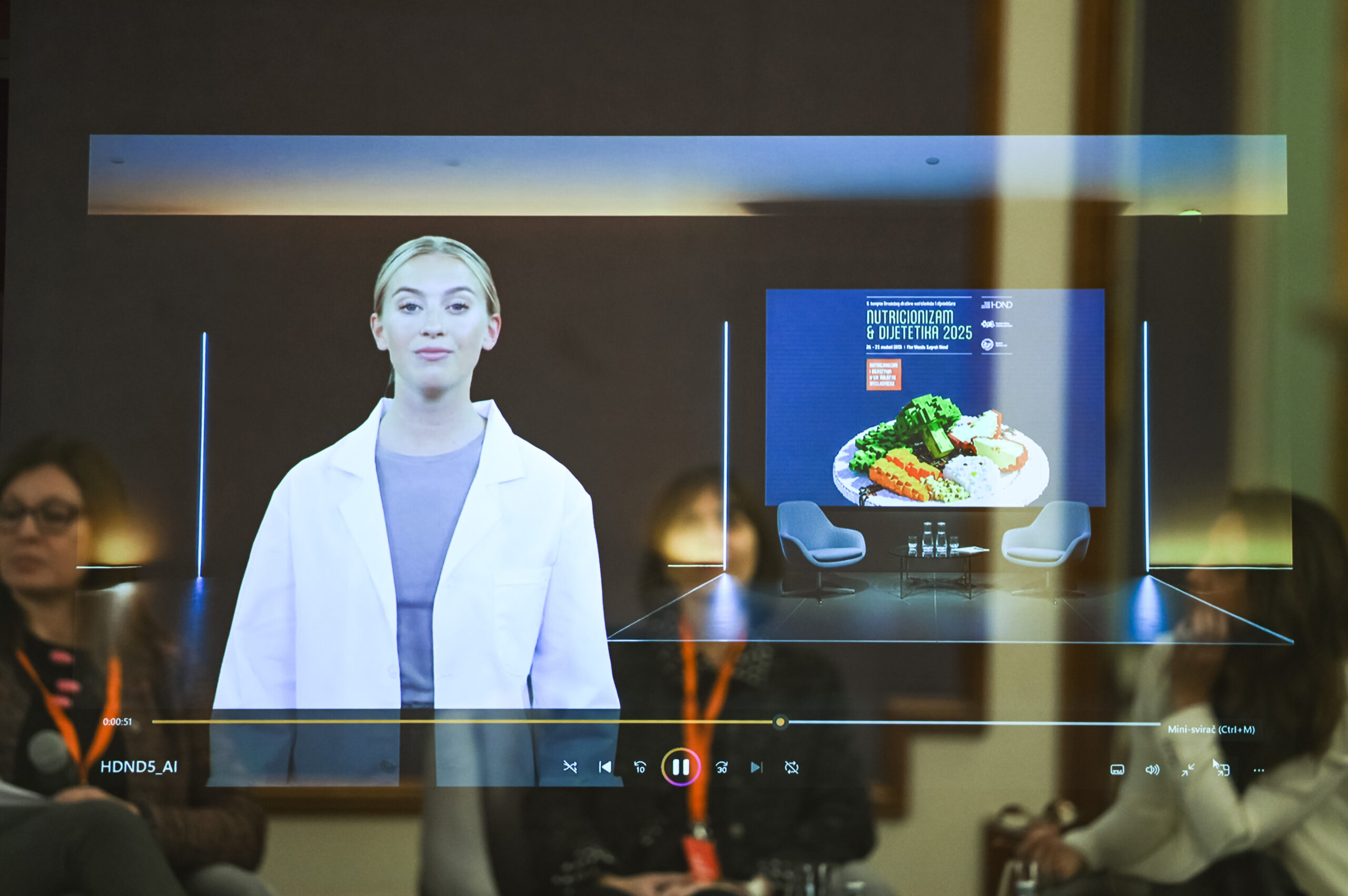 Fotografija prikazuje suvremeni konferencijski ambijent u kojem se odvija prezentacija uz pomoć hologramske ili proširene stvarnosti. Na središnjem ekranu projicirana je žena u bijelom laboratorijskom ogrtaču – vjerojatno virtualna govornica ili stručnjakinja koja sudjeluje na daljinu. Iza nje se nalazi digitalni prikaz tanjura s hranom izrađenom od šarenih građevnih kockica, što simbolizira prehranu, strukturu i znanstveni pristup dijetetici. Na pozornici su postavljene dvije stolice i stol, što sugerira da je dio programa predviđen za panel raspravu ili intervju. U prvom planu vidimo nekoliko sudionika koji pažljivo prate izlaganje. Na ekranu je istaknut naziv događaja: "Nutricionizam & Dijetetika 2025", s datumom 16. – 17. siječnja 2025. i lokacijom Hotel Esplanade, Zagreb, što potvrđuje da je riječ o stručnom skupu visoke razine. Prikazani elementi poput natpisa "HDND5_AI" i "Mini-svirka (Ctrl+M)" dodaju dozu futurističkog i interaktivnog karaktera događaju. Cjelokupna scena odiše inovativnošću, znanstvenim entuzijazmom i profesionalnim pristupom prehrambenim temama, uz naglasak na tehnologiju i multidisciplinarnu suradnju.