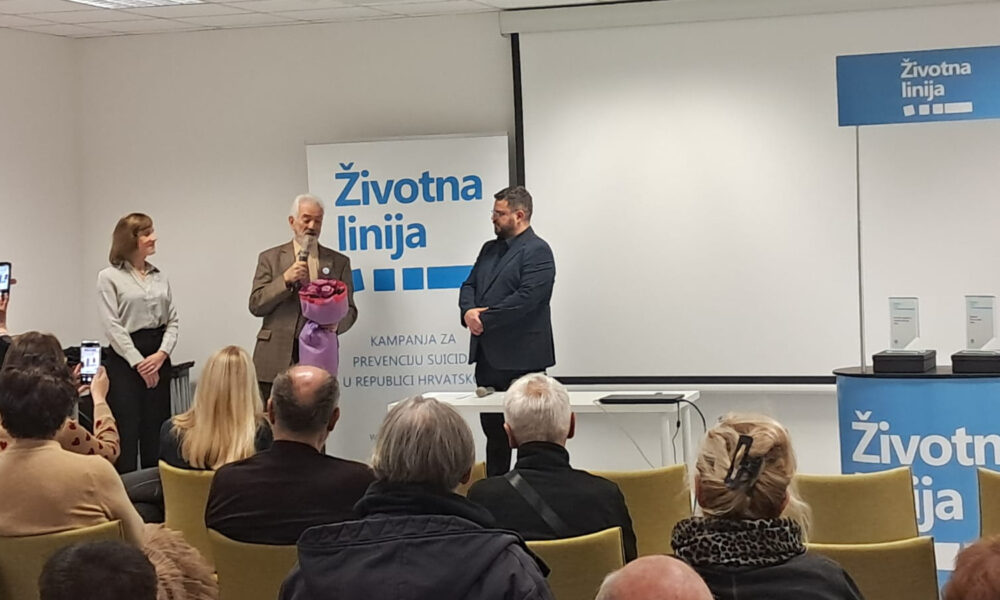 Govornik drži buket cvijeća i govori u mikrofon pred publikom, uz njega stoje dvije osobe, a u pozadini je pano s natpisom „Životna linija – kampanja za prevenciju suicida u Republici Hrvatskoj“.