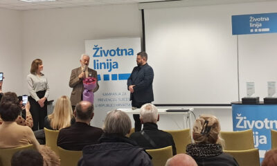 Govornik drži buket cvijeća i govori u mikrofon pred publikom, uz njega stoje dvije osobe, a u pozadini je pano s natpisom „Životna linija – kampanja za prevenciju suicida u Republici Hrvatskoj“.