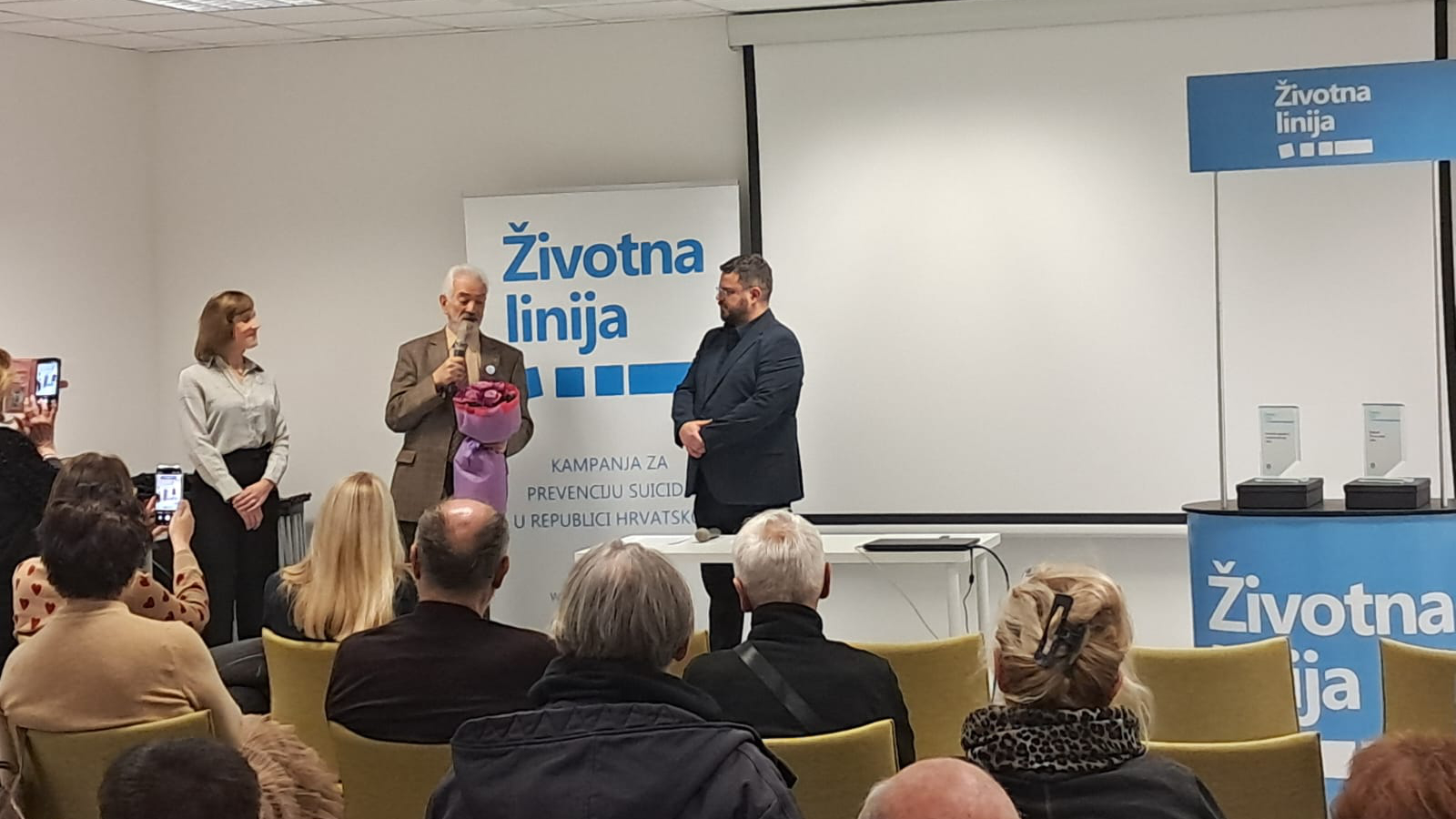 Govornik drži buket cvijeća i govori u mikrofon pred publikom, uz njega stoje dvije osobe, a u pozadini je pano s natpisom „Životna linija – kampanja za prevenciju suicida u Republici Hrvatskoj“.