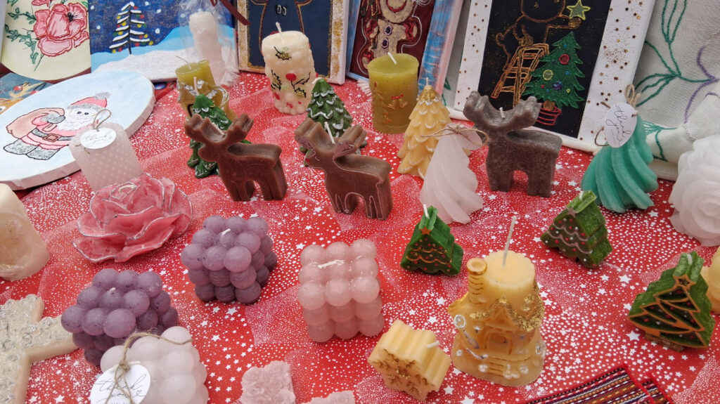 Stol prekriven crvenim stolnjakom s izloženim ručno rađenim božićnim svijećama raznih oblika i boja, uključujući jelke, sobove, anđele i kuglaste svijeće, postavljene kao dio božićnog sajma.