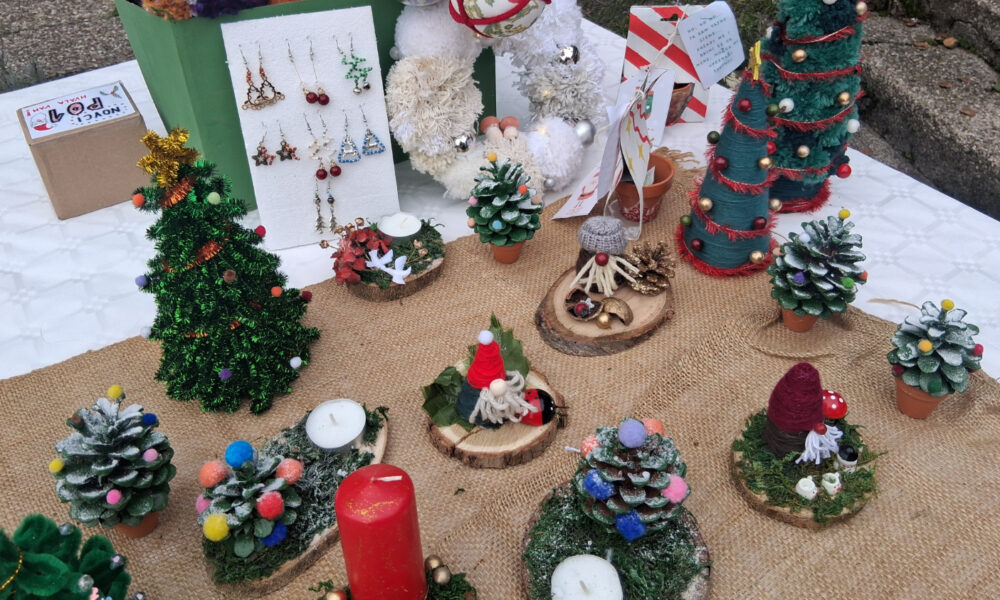 Izložbeni stol na otvorenom s božićnim rukotvorinama: male ukrasne jelke, svijeće, figurice patuljaka, nakit i prirodni ukrasi izrađeni od šišarki i drva, raspoređeni na podlozi od jute.