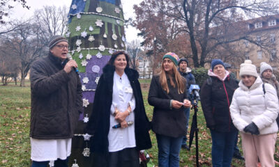 Skupina ljudi stoji u parku pored velikog božićnog drvca prekrivenog šarenim pletenim i kukičanim ukrasima. Muškarac lijevo govori u mikrofon, dok dvije žene uz njega stoje i slušaju – jedna u bijeloj kuti s tamnim kaputom, druga u zimskom kaputu i pletenoj kapi. Iza njih stoji nekoliko osoba u toploj zimskoj odjeći. Pozadina prikazuje drveće bez lišća i obližnje zgrade. Atmosfera je svečana i zimskog ugođaja.