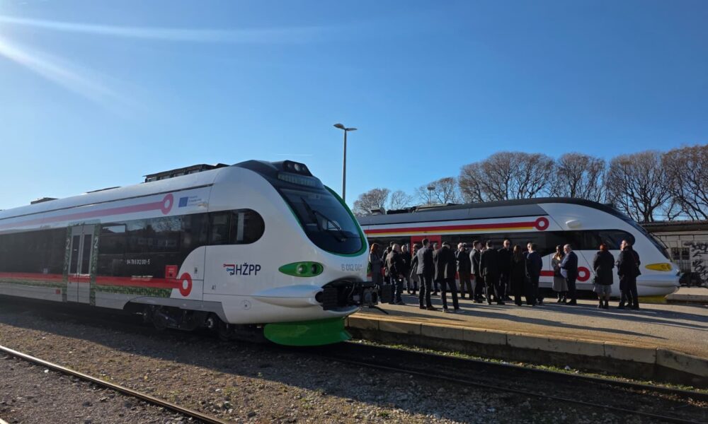 Na fotografiji se nalaze dva moderna putnička vlaka na peronu željezničke stanice, pod vedrim plavim nebom. Lijevi vlak nosi oznaku HŽPP (Hrvatske željeznice – Putnički prijevoz) i ima bijelo tijelo s zelenim i crvenim detaljima. Desni vlak ima sličan dizajn, ali s žutim i crvenim naglascima. Na peronu se okupila skupina ljudi, većinom u svečanom ili poslovnom odijevanju, što sugerira da se radi o službenom događaju — moguće predstavljanju novih vlakova ili svečanom otvorenju. Sunčeva svjetlost jasno osvjetljava prizor, a u pozadini se vide stabla bez lišća, što upućuje na zimsko ili rano proljetno razdoblje. Vidljivi natpisi na vlaku uključuju: HŽPP 91 78 6120 204-5 A1 6 012 004 Cjelokupna atmosfera fotografije odaje dojam napretka u javnom prijevozu i zajedničkog slavlja, uz naglasak na modernizaciju i uključivanje zajednice.