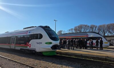 Na fotografiji se nalaze dva moderna putnička vlaka na peronu željezničke stanice, pod vedrim plavim nebom. Lijevi vlak nosi oznaku HŽPP (Hrvatske željeznice – Putnički prijevoz) i ima bijelo tijelo s zelenim i crvenim detaljima. Desni vlak ima sličan dizajn, ali s žutim i crvenim naglascima. Na peronu se okupila skupina ljudi, većinom u svečanom ili poslovnom odijevanju, što sugerira da se radi o službenom događaju — moguće predstavljanju novih vlakova ili svečanom otvorenju. Sunčeva svjetlost jasno osvjetljava prizor, a u pozadini se vide stabla bez lišća, što upućuje na zimsko ili rano proljetno razdoblje. Vidljivi natpisi na vlaku uključuju: HŽPP 91 78 6120 204-5 A1 6 012 004 Cjelokupna atmosfera fotografije odaje dojam napretka u javnom prijevozu i zajedničkog slavlja, uz naglasak na modernizaciju i uključivanje zajednice.