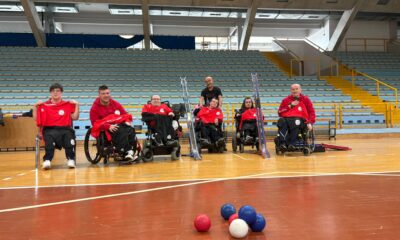 Fotografija prikazuje nekoliko ljudi u invalidskim kolicima koji su obučeni u crvene dresove, kako igraju bocciu.