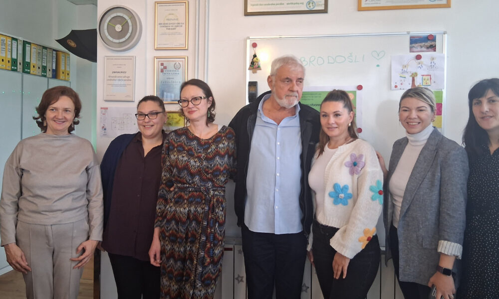 Sedam osoba stoji zajedno u uredu ispred ploče na kojoj je napisano „DOBRODOŠLI“. Među njima su četiri žene s lijeve strane, muškarac u sredini i još dvije žene desno. Na zidu iza njih vise zahvalnice i potvrde, a na ploči su dječji crteži i obavijesti. Svi se smiješe i stoje blizu jedni drugih, kao na prijateljskom ili radnom posjetu.