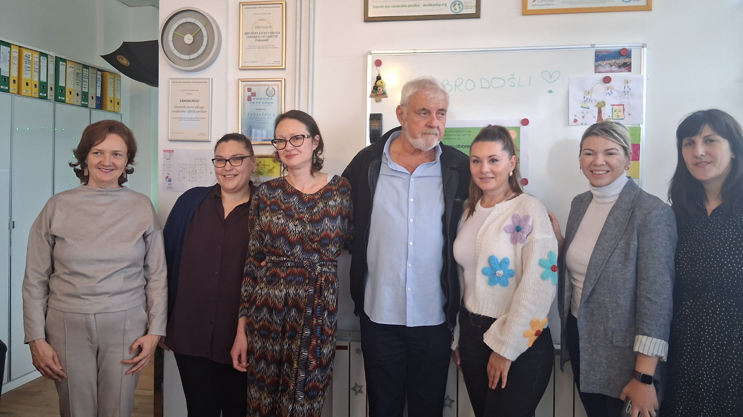 Sedam osoba stoji zajedno u uredu ispred ploče na kojoj je napisano „DOBRODOŠLI“. Među njima su četiri žene s lijeve strane, muškarac u sredini i još dvije žene desno. Na zidu iza njih vise zahvalnice i potvrde, a na ploči su dječji crteži i obavijesti. Svi se smiješe i stoje blizu jedni drugih, kao na prijateljskom ili radnom posjetu.