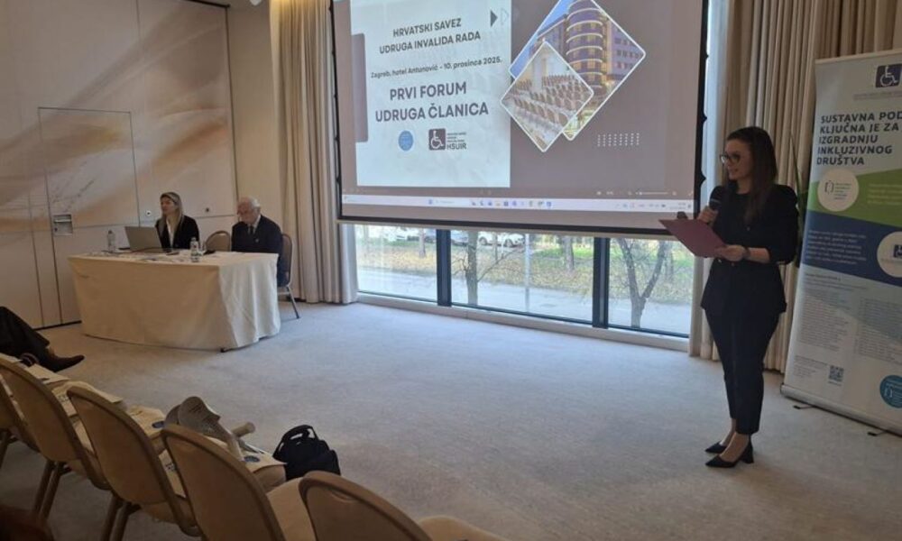Fotografija prikazuje konferencijsku dvoranu ispunjenu dnevnim svjetlom, zahvaljujući velikim prozorima koji prostoru daju otvorenost i toplinu. U prvom planu nalaze se tri osobe: Jedna osoba stoji i govori u mikrofon, držeći fascikl – vjerojatno iznosi uvodnu riječ ili prezentira temu. Dvoje sudionika sjede za stolom, ispred njih su prijenosno računalo i dokumenti, što sugerira aktivno sudjelovanje u programu. U pozadini se vidi projekcijsko platno s naslovnim slajdom prezentacije: „HRVATSKI SAVEZ UDRUGA INVALIDA RADA – PRVI FORUM UDRUGA ČLANICA“, održan 10. prosinca 2025. u hotelu Antunović, Zagreb. Desno od platna stoji vertikalni banner s porukom: „Sustavna podrška ključna je za izgradnju inkluzivnog društva“ – snažna izjava koja sažima duh događaja.