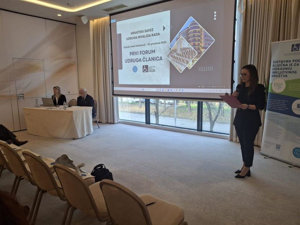 Fotografija prikazuje konferencijsku dvoranu ispunjenu dnevnim svjetlom, zahvaljujući velikim prozorima koji prostoru daju otvorenost i toplinu. U prvom planu nalaze se tri osobe: Jedna osoba stoji i govori u mikrofon, držeći fascikl – vjerojatno iznosi uvodnu riječ ili prezentira temu. Dvoje sudionika sjede za stolom, ispred njih su prijenosno računalo i dokumenti, što sugerira aktivno sudjelovanje u programu. U pozadini se vidi projekcijsko platno s naslovnim slajdom prezentacije: „HRVATSKI SAVEZ UDRUGA INVALIDA RADA – PRVI FORUM UDRUGA ČLANICA“, održan 10. prosinca 2025. u hotelu Antunović, Zagreb. Desno od platna stoji vertikalni banner s porukom: „Sustavna podrška ključna je za izgradnju inkluzivnog društva“ – snažna izjava koja sažima duh događaja.