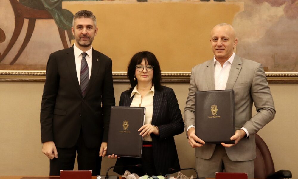 Na fotografiji su tri osobe koje stoje jedna uz drugu u formalnom okruženju, vjerojatno tijekom svečanog potpisivanja ili službenog događaja. Svi su odjeveni u poslovnu odjeću. Osobe s lijeve i desne strane drže crne mape sa zlatnim grbom i natpisom „Grad Dubrovnik“, što upućuje na povezanost s gradskom upravom Dubrovnika. Središnja osoba također drži sličnu mapu. Ispred njih se nalazi stol s mikrofonima i ukrasnim predmetom zamotanim u prozirnu foliju – moguće je da se radi o poklonu ili simboličnom predmetu vezanom uz ceremoniju. U pozadini je velika uokvirena slika klasičnog stila, što dodatno naglašava svečanu atmosferu. Ova fotografija vjerojatno dokumentira službeno partnerstvo, sporazum ili događaj od značaja za Grad Dubrovnik.