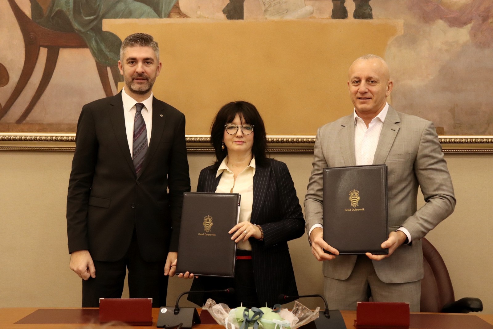 Na fotografiji su tri osobe koje stoje jedna uz drugu u formalnom okruženju, vjerojatno tijekom svečanog potpisivanja ili službenog događaja. Svi su odjeveni u poslovnu odjeću. Osobe s lijeve i desne strane drže crne mape sa zlatnim grbom i natpisom „Grad Dubrovnik“, što upućuje na povezanost s gradskom upravom Dubrovnika. Središnja osoba također drži sličnu mapu. Ispred njih se nalazi stol s mikrofonima i ukrasnim predmetom zamotanim u prozirnu foliju – moguće je da se radi o poklonu ili simboličnom predmetu vezanom uz ceremoniju. U pozadini je velika uokvirena slika klasičnog stila, što dodatno naglašava svečanu atmosferu. Ova fotografija vjerojatno dokumentira službeno partnerstvo, sporazum ili događaj od značaja za Grad Dubrovnik.