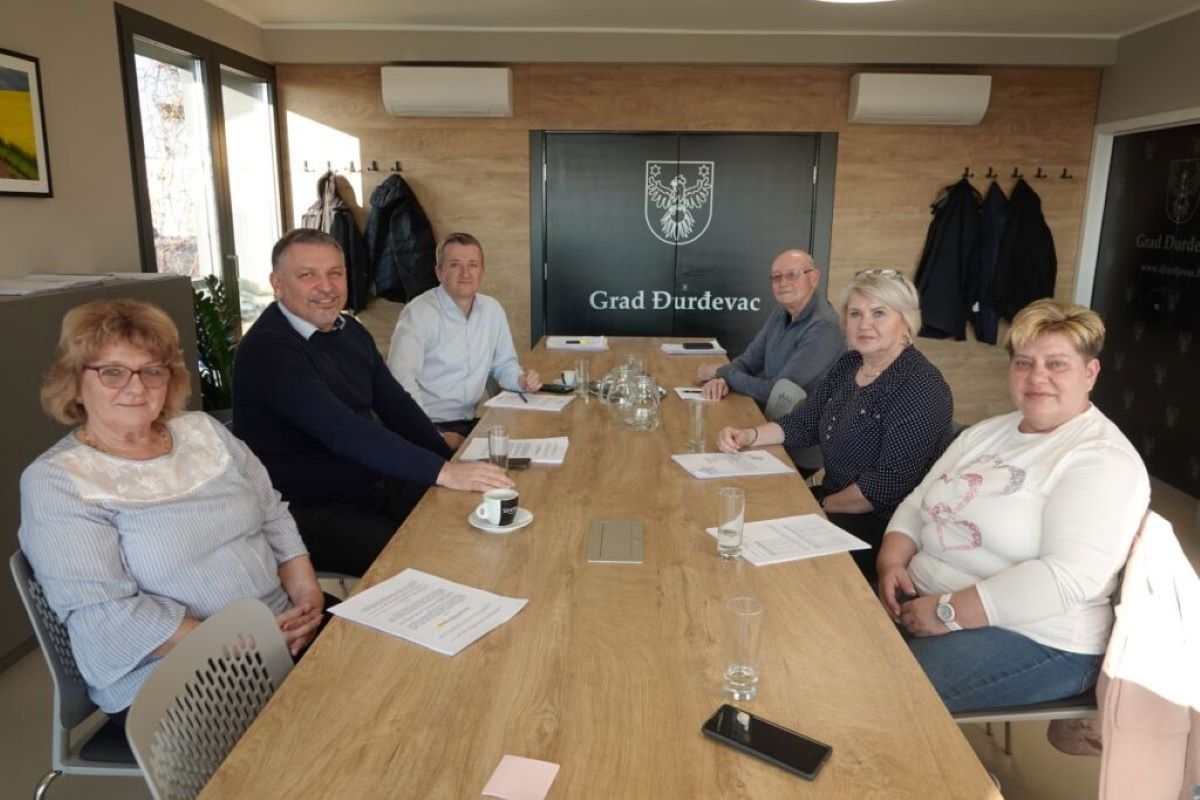 Na fotografiji je prikazano šest osoba koje sjede oko pravokutnog drvenog konferencijskog stola u modernoj prostoriji za sastanke. Svi sudionici su odjeveni formalno, a na stolu se nalaze dokumenti, kemijske olovke i čaše s vodom, što upućuje na službeni ili administrativni sastanak. U pozadini se vidi crna ploča s grbom i natpisom „Grad Đurđevac“, što sugerira da se sastanak odvija u prostorijama gradske uprave Đurđevca. Na zidu su vješalice s kaputima, a prostorija je dobro osvijetljena prirodnim svjetlom koje dolazi kroz velika prozorska stakla. Ova scena prikladna je za kontekste vezane uz lokalnu samoupravu, građansku participaciju, službene sastanke ili upravne procese.