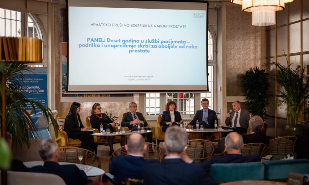 Fotografija prikazuje panel-diskusiju održanu u elegantnoj dvorani hotela Amadria Park Hotel Capital u Zagrebu. Na pozornici sjedi šest sudionika, raspoređenih u polukrugu, aktivno sudjelujući u razgovoru. Atmosfera je profesionalna i srdačna, s naglaskom na razmjenu iskustava i ideja. Publika je smještena ispred pozornice, pažljivo prati izlaganja, što dodatno naglašava važnost teme. U pozadini se nalazi veliki ekran s natpisom: HRVATSKO DRUŠTVO BOLESNIKA S RAKOM PROSTATE PANEL: Deset godina u službi pacijenata – podrška i unapređenje skrbi za oboljele od raka prostate Amadria Park Hotel Capital, Zagreb, prosinac 2023. Prostor je bogato osvijetljen, s kristalnim lusterima i dekorativnim biljkama koje doprinose toplini i svečanosti ambijenta. Vizualno, fotografija odiše dostojanstvom, zajedništvom i predanošću zdravstvenoj skrbi.