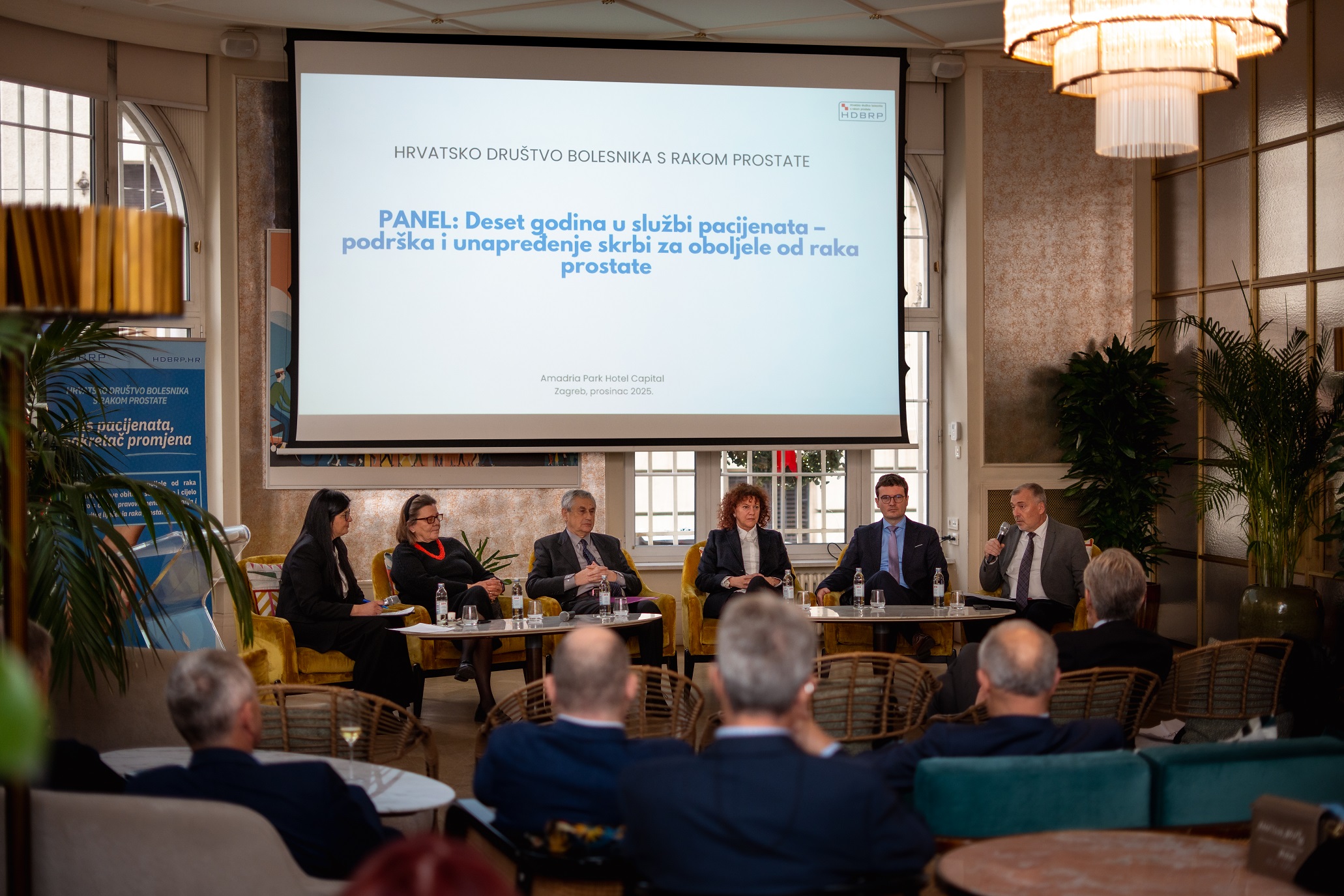 Fotografija prikazuje panel-diskusiju održanu u elegantnoj dvorani hotela Amadria Park Hotel Capital u Zagrebu. Na pozornici sjedi šest sudionika, raspoređenih u polukrugu, aktivno sudjelujući u razgovoru. Atmosfera je profesionalna i srdačna, s naglaskom na razmjenu iskustava i ideja. Publika je smještena ispred pozornice, pažljivo prati izlaganja, što dodatno naglašava važnost teme. U pozadini se nalazi veliki ekran s natpisom: HRVATSKO DRUŠTVO BOLESNIKA S RAKOM PROSTATE PANEL: Deset godina u službi pacijenata – podrška i unapređenje skrbi za oboljele od raka prostate Amadria Park Hotel Capital, Zagreb, prosinac 2023. Prostor je bogato osvijetljen, s kristalnim lusterima i dekorativnim biljkama koje doprinose toplini i svečanosti ambijenta. Vizualno, fotografija odiše dostojanstvom, zajedništvom i predanošću zdravstvenoj skrbi.