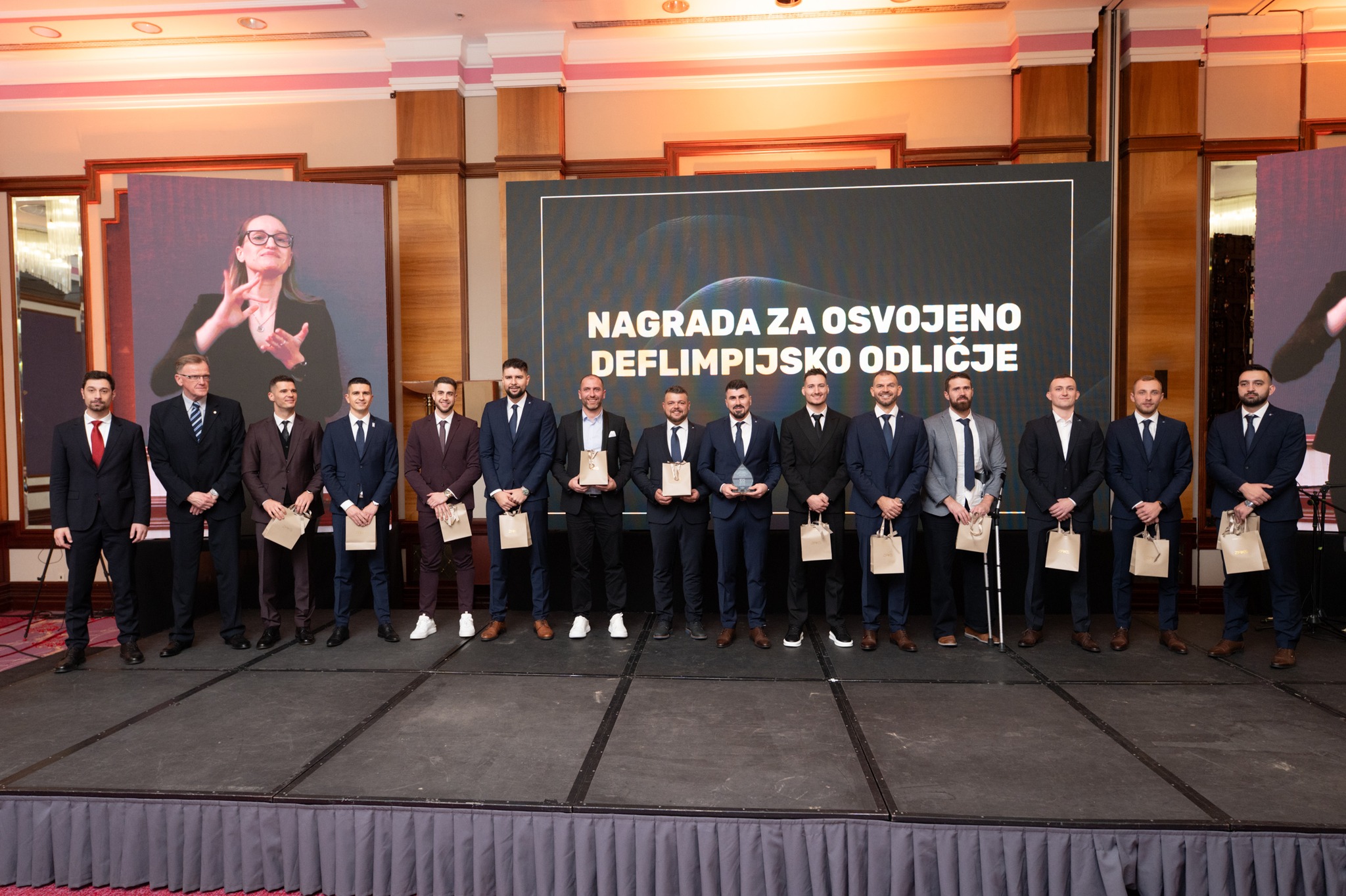 Fotografija prikazuje više ljudi kako stoje u odjelima na pozornici s vrećicama u rukama. Iza njih stoji natpis 'nagrada za osvojeno deflimpijsko odličje'.