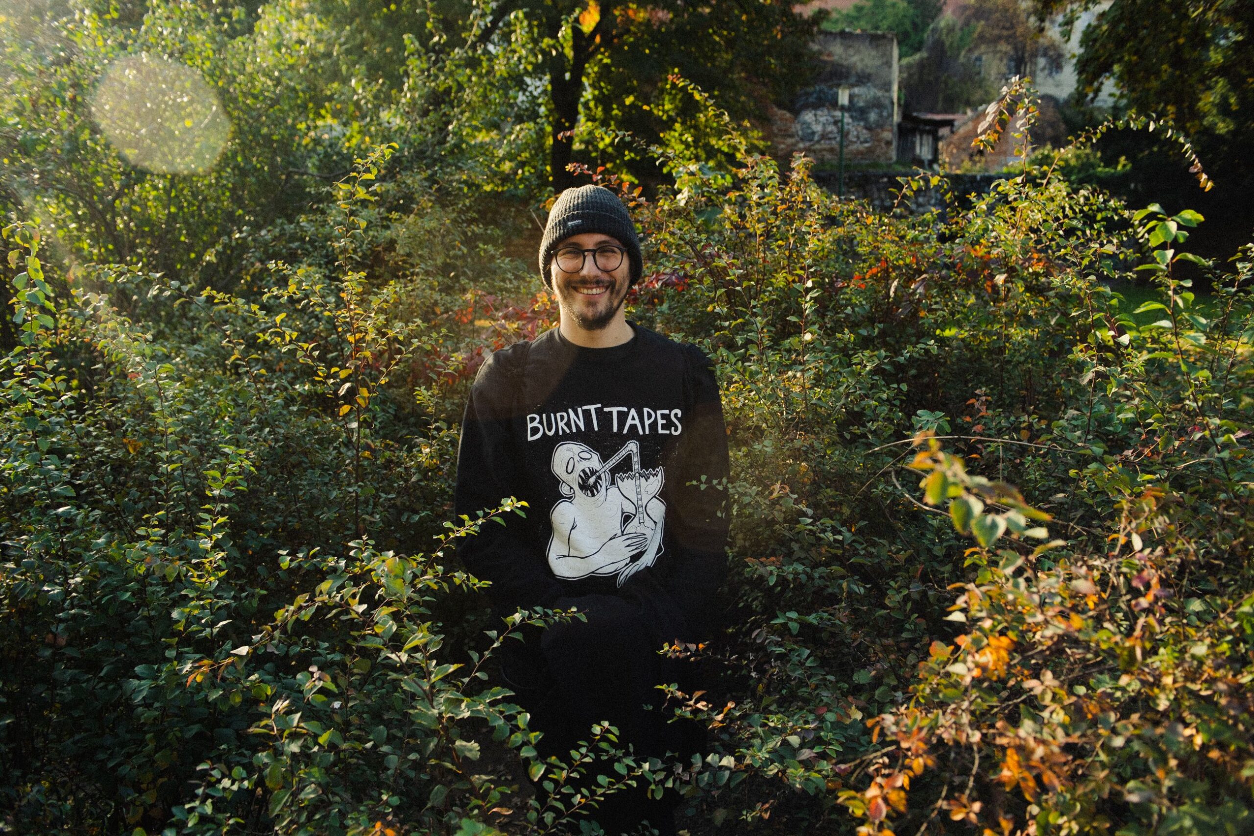 Osoba stoji nasmiješena usred gustog zelenog grmlja u sunčanoj šumskoj okolini. Nosí crnu kapu, naočale i crnu majicu s grafičkim natpisom “Burnt Tapes”. Sunčeve zrake stvaraju tople odbljeske u kadru, a u pozadini se naziru stabla i dijelovi zgrada.