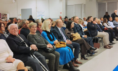 Velika skupina ljudi sjedi u nekoliko redova na svečanosti ili javnom događaju u dvorani. U prvom redu su starije osobe, parovi i pojedinci u svečanoj odjeći, neki s pomagalima poput štaka. U pozadini se vidi još publike, banner udruge i stolovi s materijalima. Atmosfera je formalna i svečana.