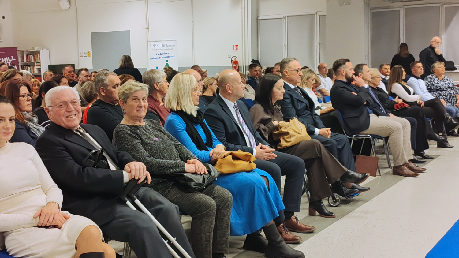 Velika skupina ljudi sjedi u nekoliko redova na svečanosti ili javnom događaju u dvorani. U prvom redu su starije osobe, parovi i pojedinci u svečanoj odjeći, neki s pomagalima poput štaka. U pozadini se vidi još publike, banner udruge i stolovi s materijalima. Atmosfera je formalna i svečana.