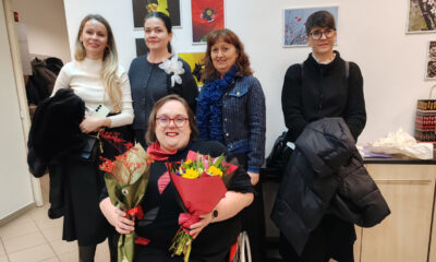 Skupina žena u zatvorenom prostoru pozira za fotografiju; u prvom planu žena u invalidskim kolicima drži dva buketa cvijeća, dok ostale stoje iza nje, uz zid s izloženim fotografijama, tijekom događanja.