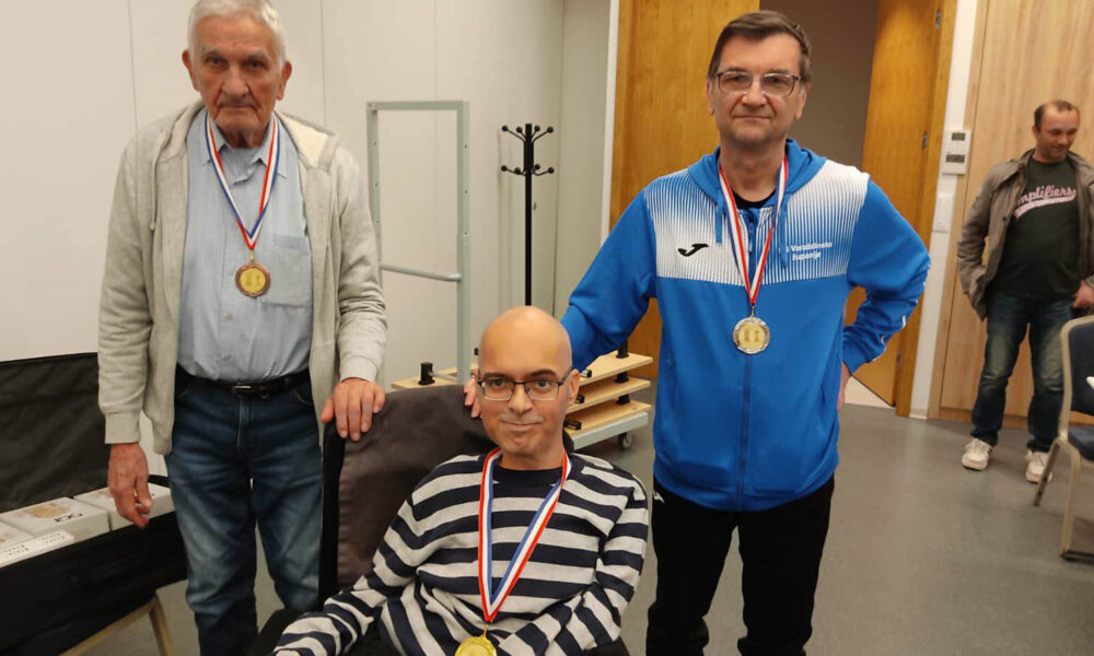 Troje muškaraca pozira s medaljama oko vrata u unutarnjem prostoru. Dvojica stoje, a jedan sjedi u invalidskim kolicima. Svi nose natjecateljske medalje i blago se smiješe prema kameri. U pozadini se vidi neutralan interijer s drvenim vratima i nekoliko komada namještaja.