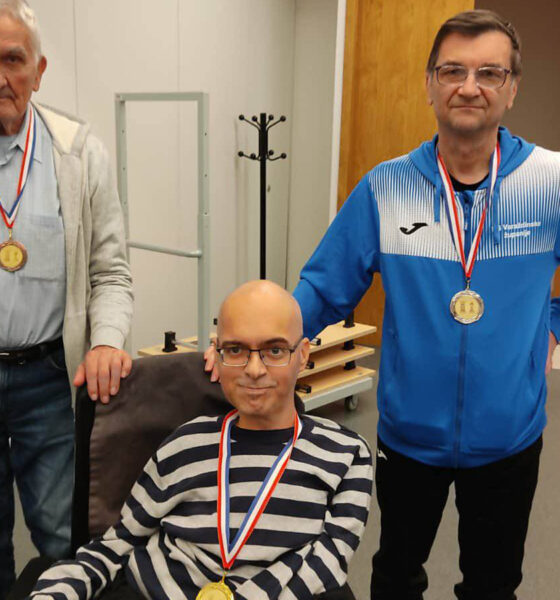 Troje muškaraca pozira s medaljama oko vrata u unutarnjem prostoru. Dvojica stoje, a jedan sjedi u invalidskim kolicima. Svi nose natjecateljske medalje i blago se smiješe prema kameri. U pozadini se vidi neutralan interijer s drvenim vratima i nekoliko komada namještaja.