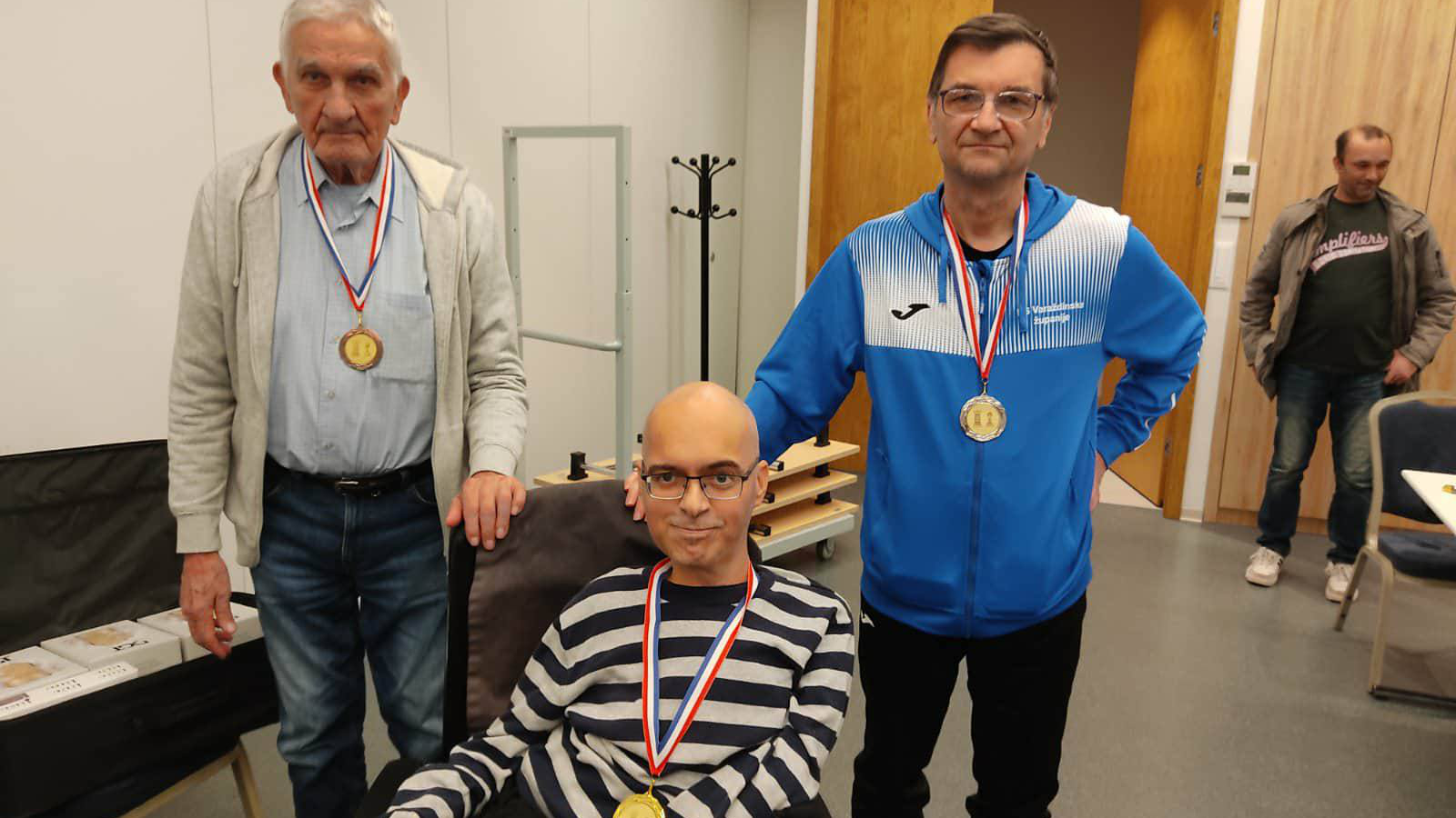 Troje muškaraca pozira s medaljama oko vrata u unutarnjem prostoru. Dvojica stoje, a jedan sjedi u invalidskim kolicima. Svi nose natjecateljske medalje i blago se smiješe prema kameri. U pozadini se vidi neutralan interijer s drvenim vratima i nekoliko komada namještaja.